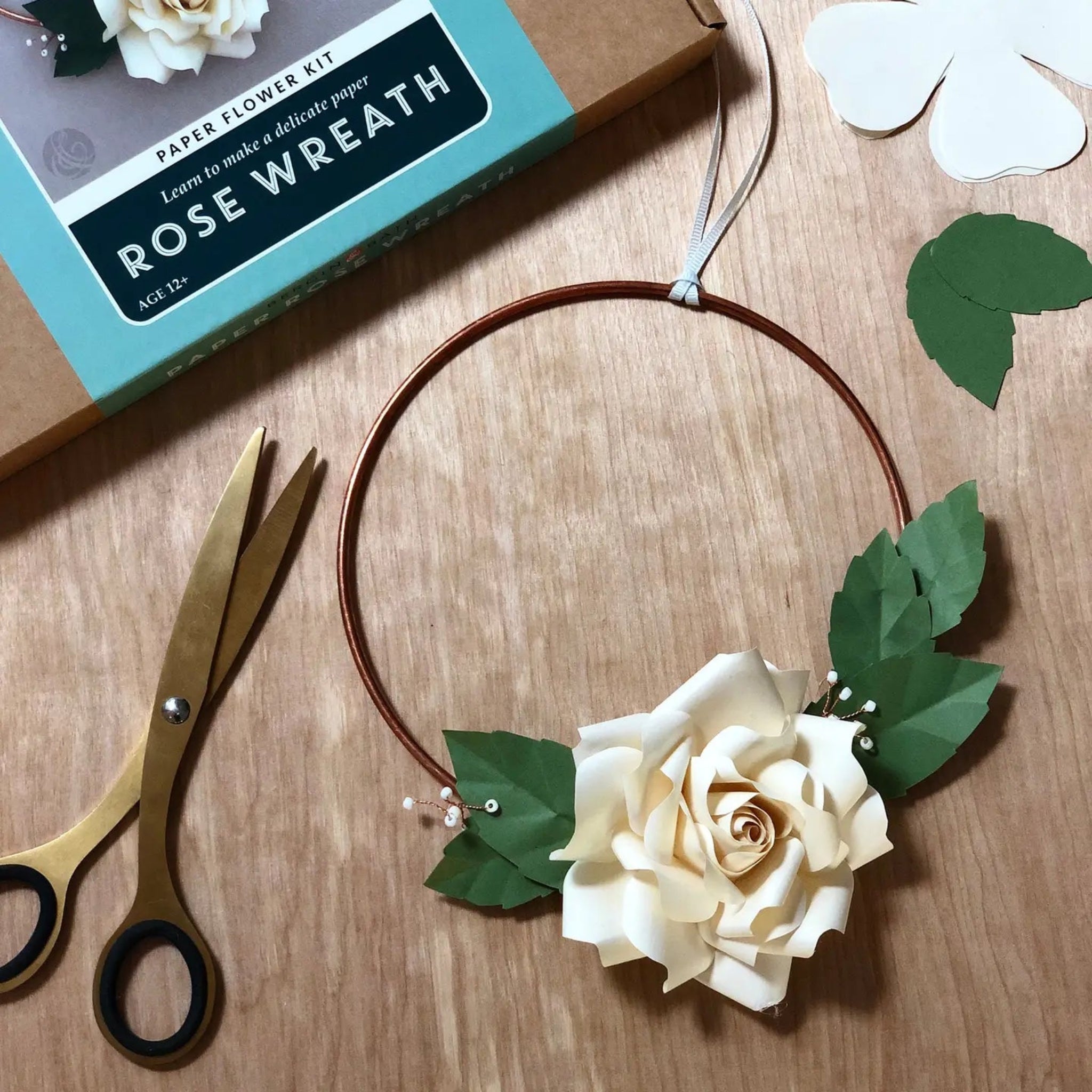 Paper flower kit with a completed rose wreath, scissors, and packaging on a wooden surface.