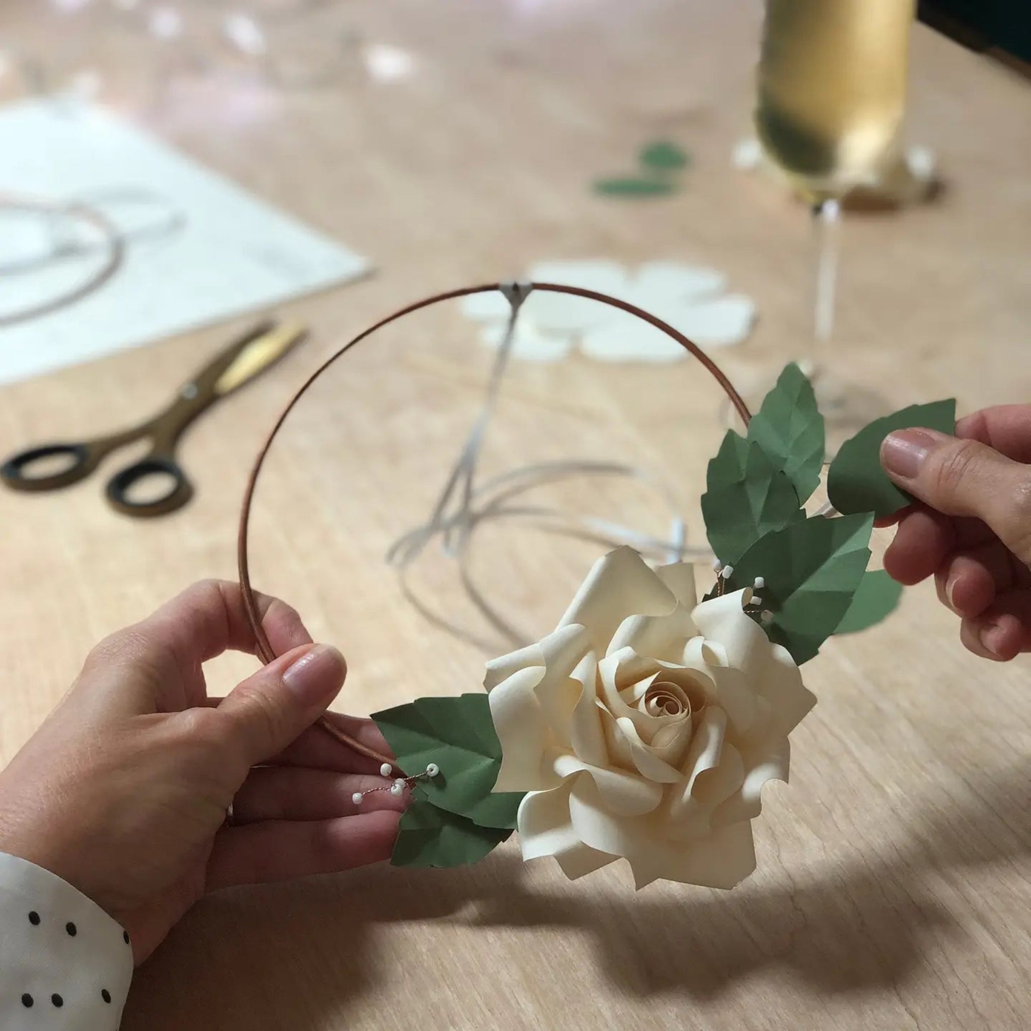 Floral headband with white roses and green leaves held by hands on a wooden surface.
