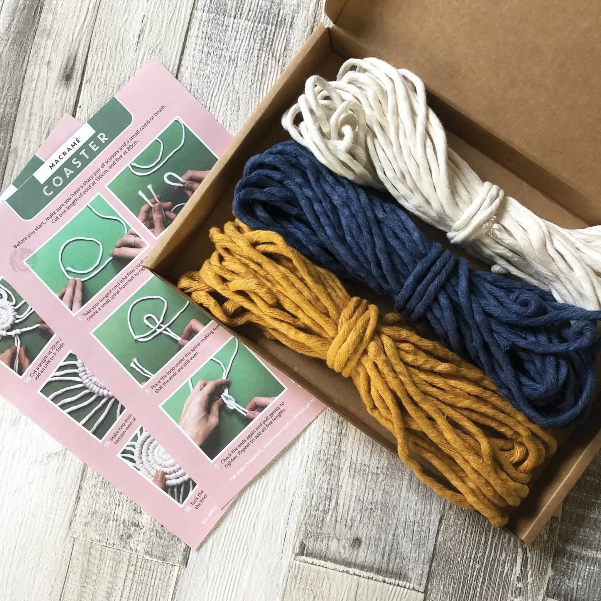 Box containing yarn in blue, yellow, and white with a pink coaster-making guide on a wooden surface.